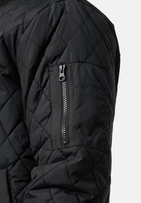 Veste matelassée noire avec un extérieur texturé, dotée d'une poche zippée sur la manche et d'un design rembourré pour l'isolation.