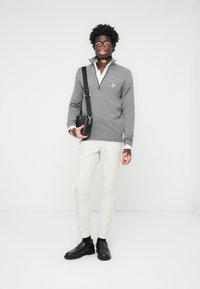 Grauer Reißverschluss-Pullover über einem weißen Hemd, weißen Hosen und schwarzen Schuhen. Das Model trägt eine schwarze Umhängetasche. Einfaches Design mit minimalen Akzenten.
