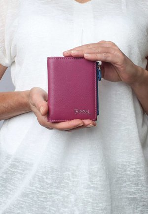 Portafoglio in pelle magenta con una texture liscia, chiusura zip e dettaglio del logo in argento. Tenuto in mano su uno sfondo di indumento chiaro e testurizzato.