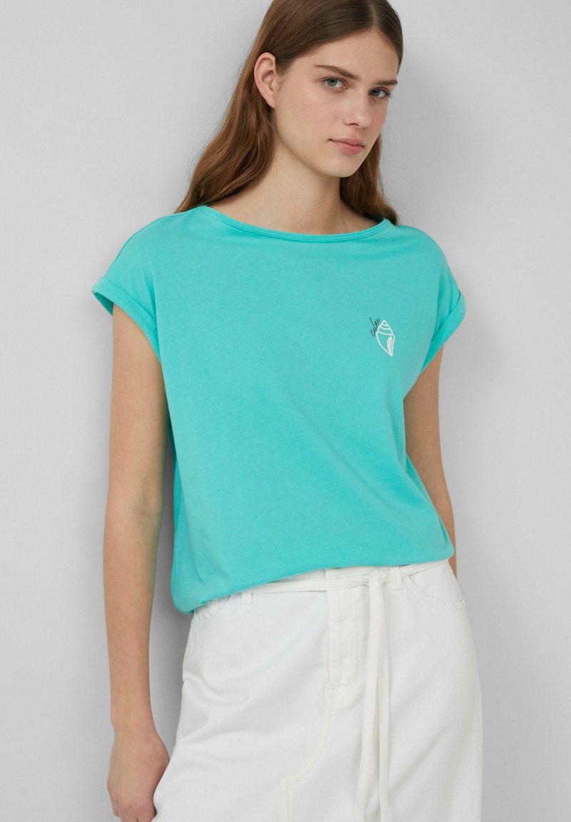 Turkusowa bawełniana koszulka z podwiniętymi rękawami i okrągłym dekoltem; na lewej stronie znajduje się mała biała grafika muszli.
