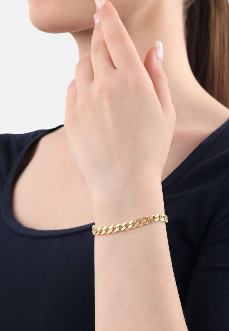 Mujer que lleva una pulsera de eslabones de oro en la muñeca, con la mano levantada cerca de su rostro, vistiendo una blusa de color azul marino oscuro.