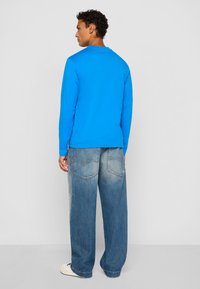 Mand står med ryggen til iført en lys blå langærmet skjorte, løse, falmede blå jeans og hvide sneakers foran en ensfarvet baggrund.
