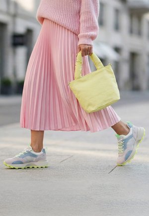 Person geht auf einer Stadtstraße und trägt einen pinken plissierten Rock, pastellfarbene Sneaker, einen pinken Pullover und hält eine gelbe Handtasche in der Hand.