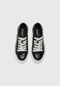 Svarta lacklädersneakers med vita snören, med texturerade accenter och en platt vit sula. Logotyper synliga på tungan och sidan.