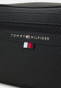 Black textured leather bag with a top zipper. Features silver "TOMMY HILFIGER" branding and a small red, white, and blue accent detail.