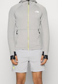 Chaqueta con capucha de color gris claro con cremallera, tela texturizada, cremallera de color amarillo brillante, detalles en negro en los puños y los laterales, llevada sobre pantalones cortos de color gris claro.