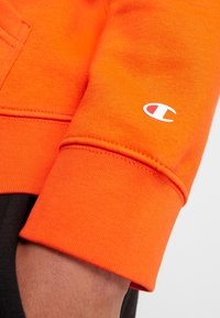 Sudadera naranja con un acabado texturizado, que presenta un logo blanco en el puño. Incluye un bolsillo y detalles acanalados en la manga.