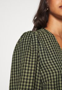 Blouse à carreaux verts et noirs avec un tissu texturé, présentant des manches bouffantes et un décolleté en V. Gros plan sur le détail de l'épaule.