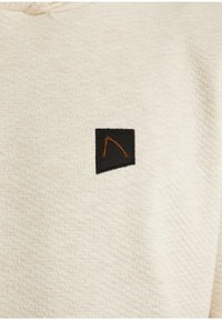 Hoodie beige clair avec tissu texturé. Présente un patch noir avec une couture orange dans un design géométrique, situé sur la poitrine.