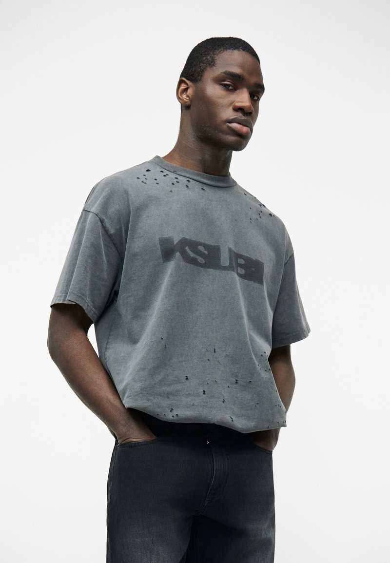 Joven con las manos en los bolsillos, vistiendo una camiseta gris descolorida con pequeños agujeros y pantalones oscuros, sobre un fondo blanco liso.