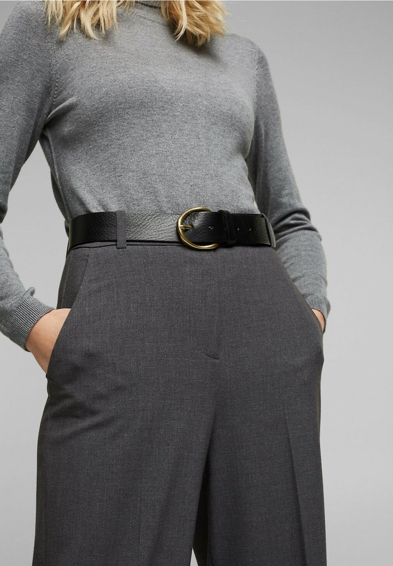 Femme portant un pull gris ajusté rentré dans un pantalon gris foncé taille haute avec une large ceinture noire à boucle dorée.