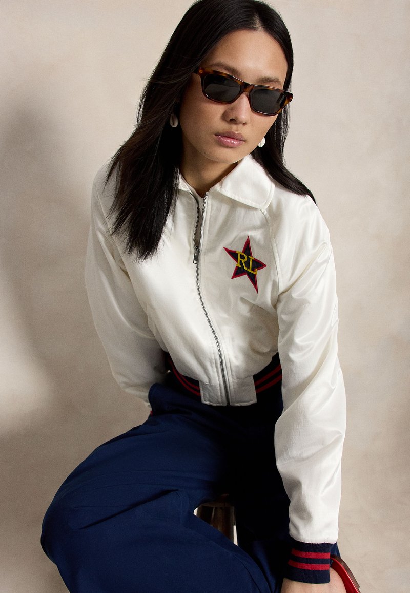 Femme aux longs cheveux foncés portant des lunettes de soleil écaille de tortue, une veste blanche zippée avec un emblème étoile, et un pantalon bleu marine assise devant un fond uni.