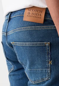 Jean en denim bleu avec une étiquette en cuir sur la ceinture arrière et des coutures contrastées sur les poches arrière.