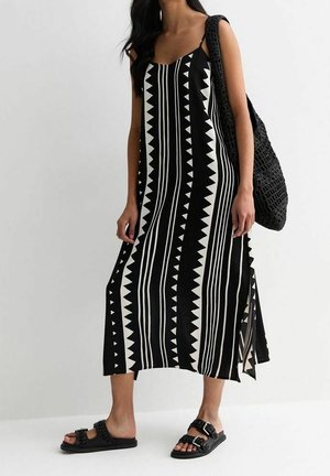 Žena v černo-bílých vzorovaných midi šatech, černých sandálech a s velkou černou pletenou taškou přes rameno.