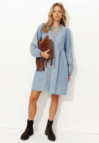 Robe en denim bleu clair avec un devant à boutons, des détails brodés, de longues manches bouffantes et une jupe évasée. Assortie à des bottines marron.