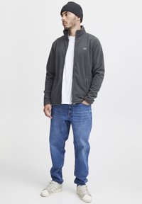 Gray fleece jacket with high collar, zippered front, two pockets, worn over a white shirt. Paired with blue jeans and white sneakers.