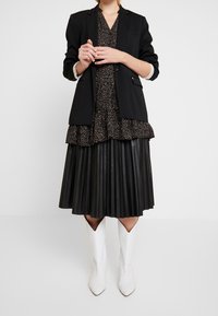 Czarna marynarka nałożona na sukienkę w grochy z falbanką, warstwą pod plisowaną czarną spódnicą. Białe, teksturowane botki za kostkę dopełniają całość.