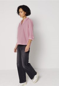 Blusa rosa chiaro con scollo a V e maniche arrotolate, abbinata a pantaloni larghi grigi scuri e sneakers bianche, indossata in modo casual.