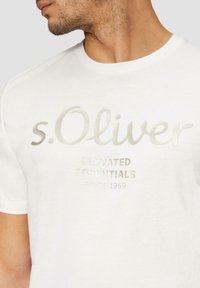 Bílý bavlněný tričko s metalickým stříbrným textem. Na přední straně je vytištěno "s.Oliver" a "ELEVATED ESSENTIALS SINCE 1969".