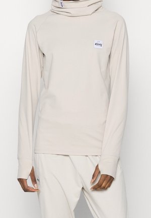 Person iført en beige ribbet langærmet top med høj hals og matchende bukser, begge med små hvide rektangulære mærkeetiketter.