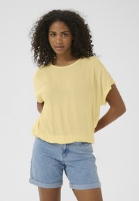 Blouse jaune surdimensionnée avec un col rond et des manches courtes, associée à un short en denim avec des ourlets retroussés et un lavage clair.