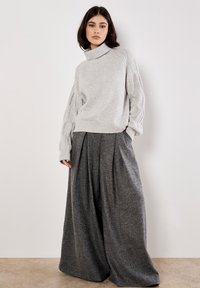 Maglione grigio chiaro con colletto alto e maniche testurizzate, abbinato a pantaloni larghi grigio scuro con pieghe frontali.