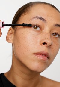 Du mascara noir est appliqué avec un pinceau. Le sujet a la peau brun clair avec des taches de rousseur, des yeux marron et des lèvres lisses. Un applicateur rose subtil est visible.