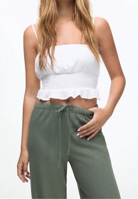 Haut court blanc smocké avec ourlet à volants, associé à un pantalon vert à cordon de serrage. Textures lisses et coupe décontractée mises en avant dans le design.