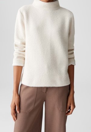 Personne portant un pull blanc texturé à manches longues et col montant, associé à un pantalon marron ample avec des poches plaquées à l'avant.