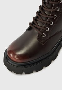 Sötétbarna bőr csizma vastag fekete lug talppal és azonos színű barna fűzőkkel, melyek fémszemeken keresztül vannak átfonva.