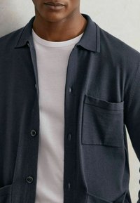 Hombre vistiendo una camisa de botón azul oscuro sobre una camiseta gris claro, cortada a la altura de los hombros y del torso medio.