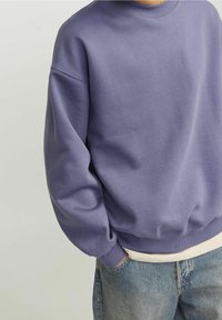Person, der einen lockeren, langärmeligen lila Sweatshirt mit gerippten Bündchen und Saum trägt, kombiniert mit blauen Jeans und einem weißen Hemd darunter.