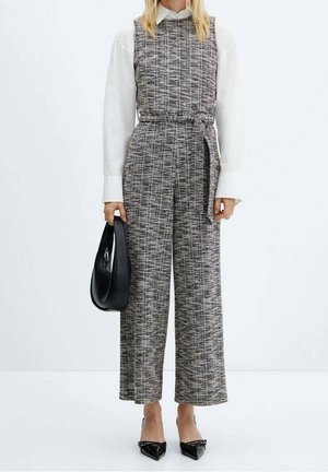 Kvinde iført grå tweed ærmeløs jumpsuit med bælte, hvid langærmet skjorte, sorte spidse hæle og holder en sort håndtaske.