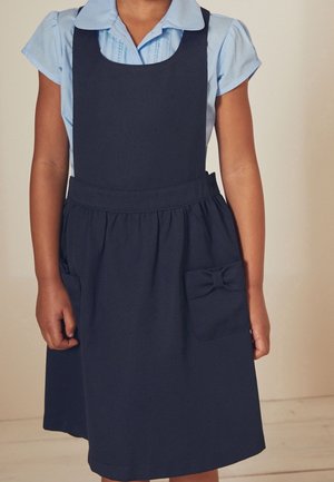 Enfant portant une chemise bleu clair à manches courtes sous une robe chasuble bleu marine avec un nœud sur la poche, debout devant un fond uni.