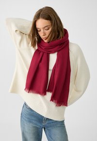 Sciarpa cremisi realizzata in morbido tessuto con bordi frangiati, avvolta attorno al collo, abbinata a un maglione crema e jeans blu.