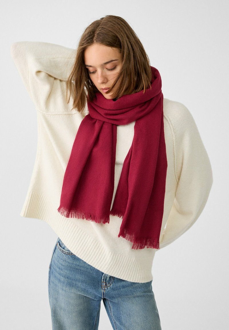 Sciarpa cremisi realizzata in morbido tessuto con bordi frangiati, avvolta attorno al collo, abbinata a un maglione crema e jeans blu.