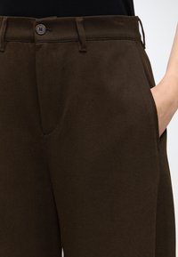 Gros plan sur un pantalon marron foncé avec passants de ceinture, fermeture par bouton et une main partiellement glissée dans la poche latérale droite.