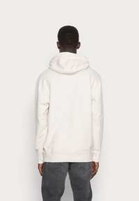 Felpa beige chiaro con cappuccio regolabile, polsini e fondo a coste, e una consistenza morbida, indossata sopra pantaloni grigi, vista da dietro.