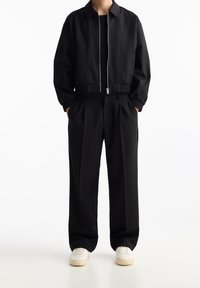 Zwarte cropped jas met ritssluiting en elastische boorden, gecombineerd met losse zwarte broek met plooien; neutraalgekleurde sneakers maken de look compleet.