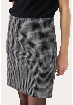 Gros plan d'une personne portant une jupe crayon taille haute à motif pied-de-poule avec un haut noir et des collants noirs translucides.