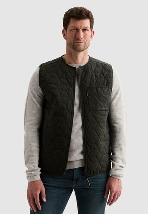 Man wearing a black quilted vest over a light gray textured long-sleeve shirt and blue jeans, facing forward against a plain background.