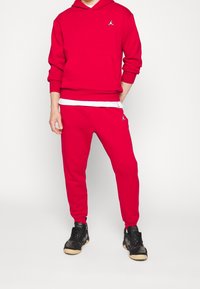 Červená mikina s kapucí a přední kapsou, spárovaná s odpovídajícími červenými tepláky s logem. Černé sportovní boty dokončují celý outfit.