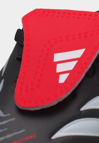 Κοντινό πλάνο ενός κόκκινου επενδυμένου λουριού με λευκό λογότυπο πάνω σε αθλητικό παπούτσι μαύρου και γκρι χρώματος, που δείχνει λεπτομερή ραφή και υφή επιφάνειας.