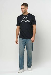 Mann iført svart t-skjorte med grafisk trykk, lyseblå jeans, hvite joggesko og sølvkjede som går mot en enkel hvit bakgrunn.