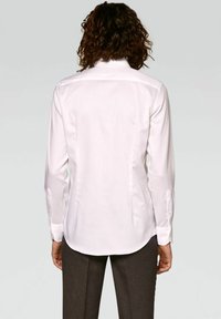 Chemise blanche à manches longues avec col pointu, tissu texturé, ourlet arrondi et poignets boutonnés, vue de dos avec un pantalon foncé.