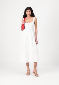 Abito midi bianco in cotone con motivi a stella ricamati e scollatura quadrata. Abbinato a sandali nudi con lacci e una borsa rossa.