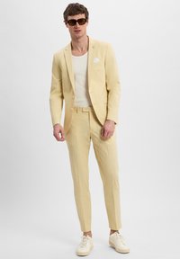 Helles gelbes Anzug mit schmaler Passform, einreihiger Jacke und passender Hose. Darunter ein weißes Tanktop, kombiniert mit braunen Sonnenbrillen und weißen Sneakers.