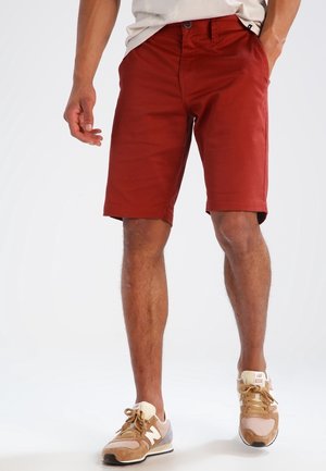 Mann trägt rostrote Shorts und beige-braune Sneakers, steht mit einer Hand in der Tasche und der anderen entspannt an der Seite.