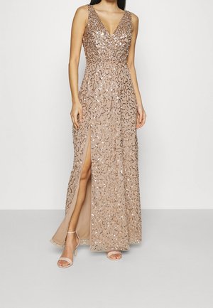 Kvinde iført en ærmeløs, V-udskæring, gulvlængde beige kjole med palietter og en frontslids op til låret, kombineret med nøgenfarvede høje hæle.