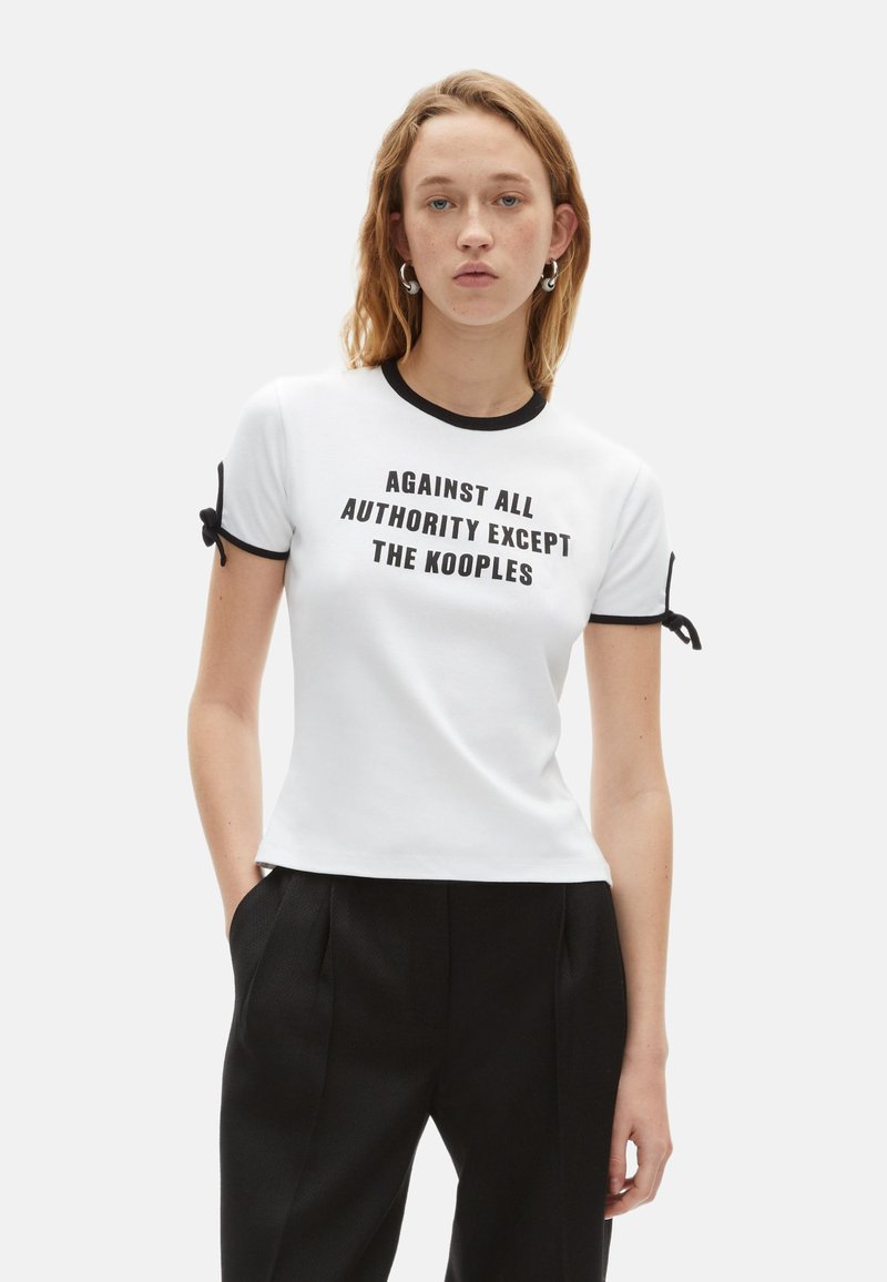 Hvid kortærmet t-shirt med sort tekst "MOD ALLE MYNDIGHEDER BORTSET FRA KOOPLES," med kontrasterende sort kant og snoredetaljer på ærmerne.
